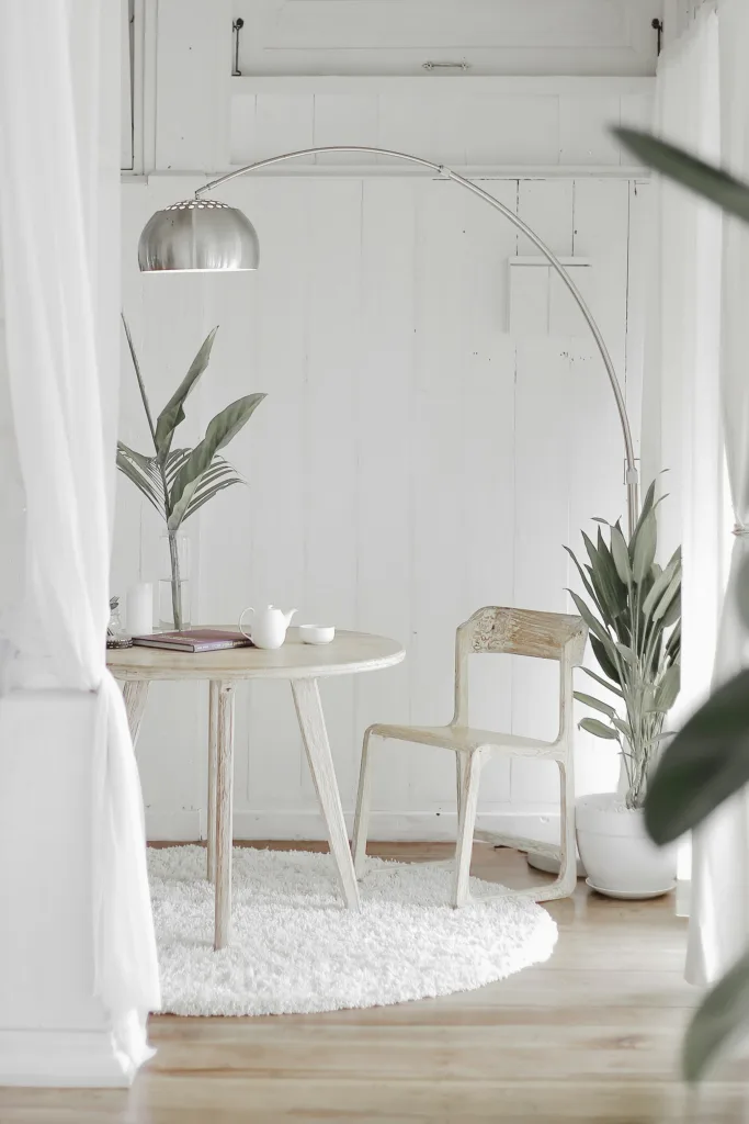 Minimalist white room with a round wooden table, arched floor lamp, chair, and potted plants on a shag rug