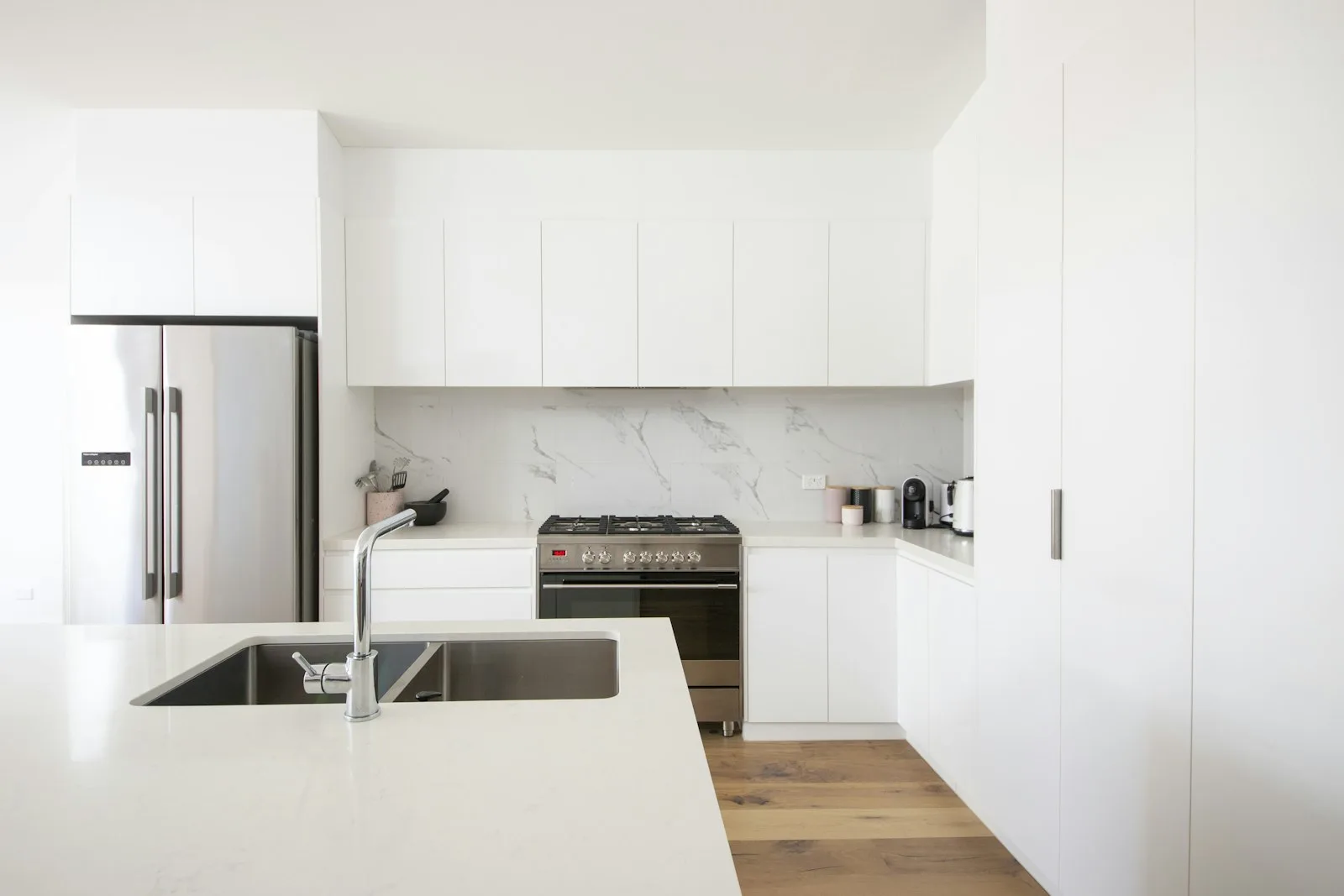 Modern white kitchen with stainless steel appliances, marble backsplash, and a large island sink in the foreground