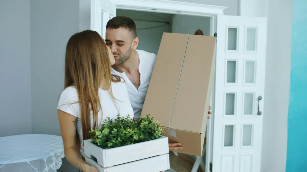 Couple kissing at a doorway while carrying moving boxes and a potted plant