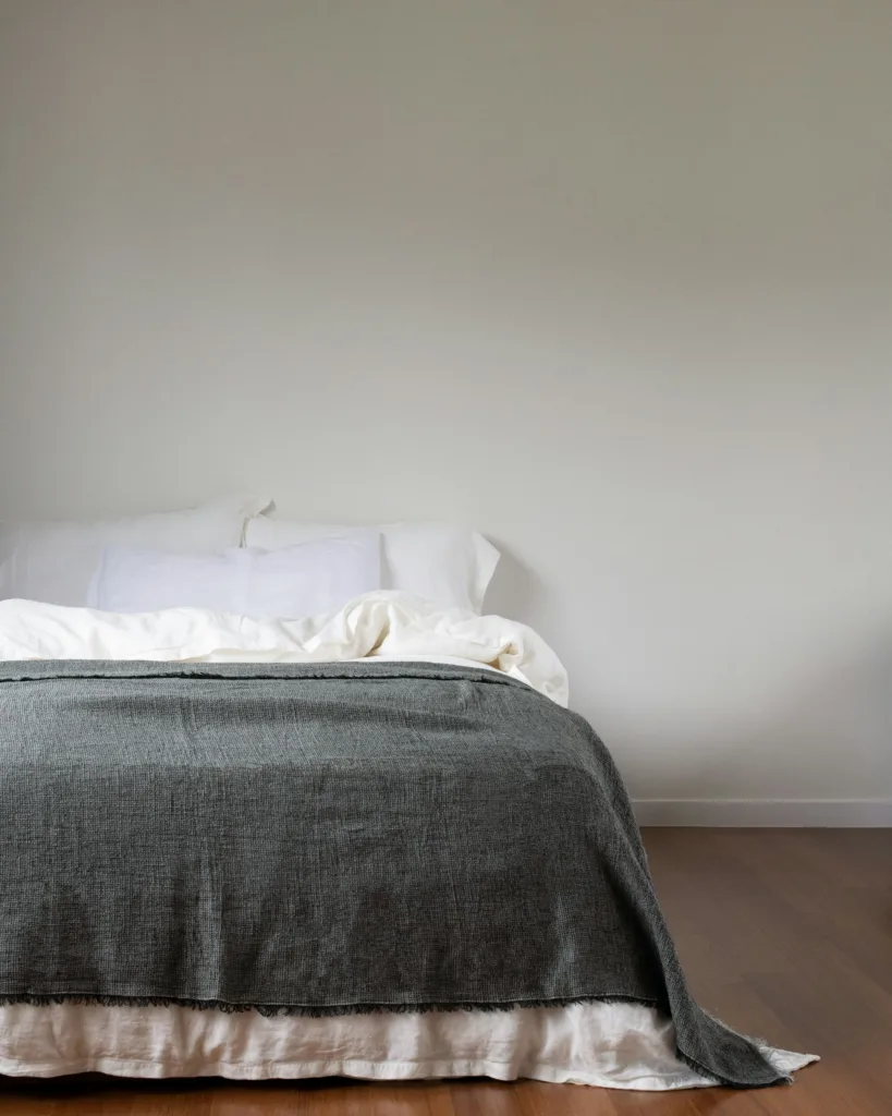 Minimalist bed with white pillows, rumpled white sheets, and a dark gray blanket against a plain wall